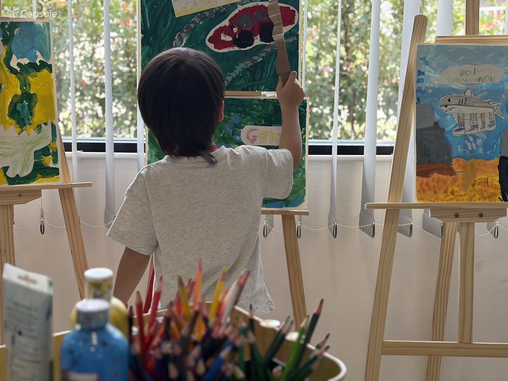 เด็กหันหลังวาดรูปที่ขาตั้งภาพในสตูดิโอ Little Capsule ระหว่างแคมป์ศิลปะ Emotion Explorers